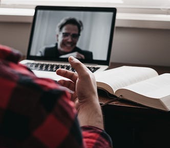 Person working at a desk with a laptop showing a male in full-screen on a video call. Next to the laptop lies an open book. The person partly in the frame holds their hand up, as if explaining something to the person on-camera.