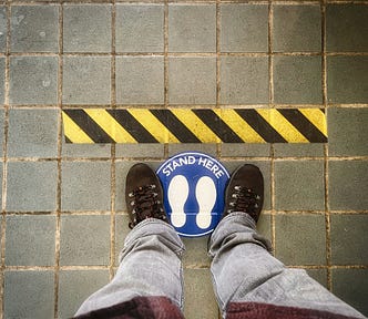 View down on feet standing on a “stand here” mark, behind a black and yellow warning line.