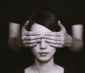Photo of a woman facing the camera. A man is standing behind her and has his hands covering her eyes.