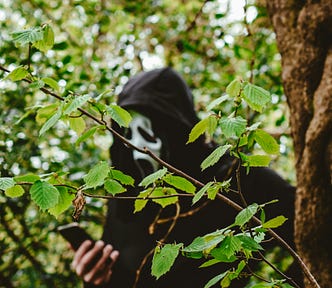 Guy in a scream mask, standing behind a tree and checking his phone
