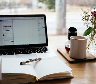 A pen, notepad, coffee cup, and a laptop on a wooden table.