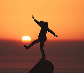 A person stands on their right foot on a rock. Their left lef extends forward at a 45 degree angle. The right hand extends upward for balance (and the left slightly downward). The sun is setting in the background, rendering the entire image an orange hue. One in five individuals in a recent study could not stand on one foot for ten seconds.