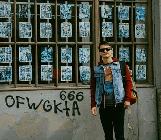 Young man wearing sunglasses, a red hoodie with design partially hidden by a denim vest, black jeans, and a backpack. He is standing next to a wall with a large window full of promotional posters. Beneath the wall is spray-painted 666 and random letters.