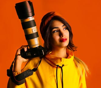 A woman in a yellow jacket and red head scarf holding a camera with a large lens in a vibrant reddish orange background