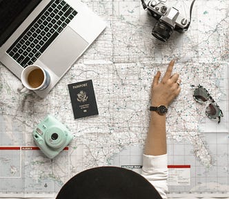 On top of a detailed map of the United States likes a laptop and mug of coffee in the top left corner, a Polaroid-style camera and passport beneath them, a fancier digital camera in the top right, and a pair of sunglasses in the middle right. At the bottom there is an individual in a white shirt and wide-brimmed black hat viewed from above, a finger pointing out a location on the map, potentially tracing out a route.