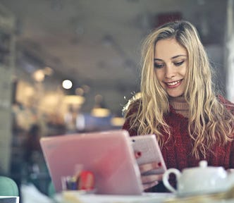 Blonde woman in red sweater working on her laptop and looking at her phone smiling with a blurred office behind her