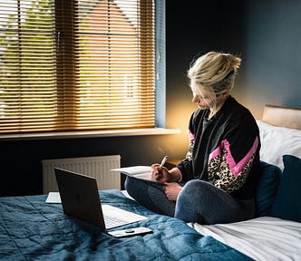 Woman writing in a notebook, sitting on her bed, with a computer in front of her.