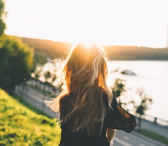 A woman stares out at the sunrise view of a boulevard by the water’s edge, a lighthouse in the distance.