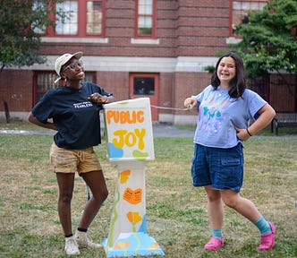 Sabrina and Ava stand with a four-foot-tall box between them on a patch of grass. They are both smiling, and Ava opens the top of the box. The box is painted with bright orange, yellow, green, and blue colors and says the words “Public Joy”.