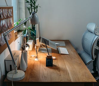 Work from home setup with wooden desk, sat directly at a small window, with minimal appliances, several small plants, a macbook on a standard, a keyboard and a mouse. On the wall there is a cork board with pinned images into it.
