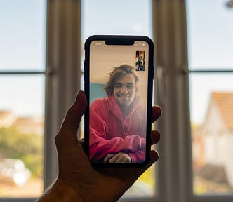 A hand holds a phone and the phone displays someone who is video chatting with the person on the other end of the hand — a blurry window and suburban scene with a house and car is in the background