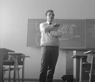 Black and white photo with a man standing in front of a black board with desks around.
