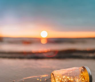 A sunset horizon view on the beach from the sand overlooking a lying glass jar with little string lights inside (that mimic the look of fireflies) and a receding wave. The similar glow of the sunset jar lights and sunset are like bulbs from the same company of lights.