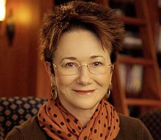 headshot of white woman with short spiky brown hair wearing orange scarf w black polkadots and rimless eyeglasses. big earrings. smiling at camera. she’s sitting in a library with a ladder for reaching books.