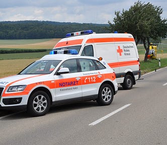 White and orange ambulance, emergency vehicle, and medi-vac helicopter pulled over to the side of the road, next to a field.