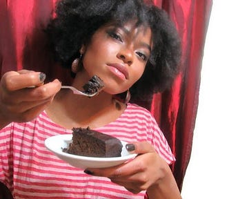 Have some? Person holding a small plate with a slice of black cake — also called Christmas cake or Caribbean fruit cake. The other hand holds a fork with a bite of the cake to share.