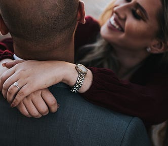 Couple locking arms around other persons neck. To the left, a black male with short hair. We see him from the back. On the right, a white female, smiling.