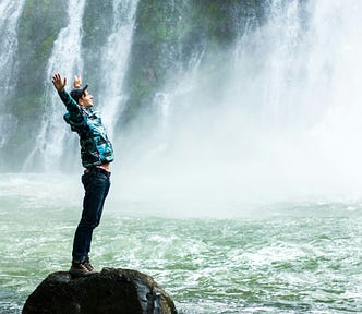 Male standing at the side of a waterfall wearing a cap, sporty clothes, arms up in the air, he looks happy.