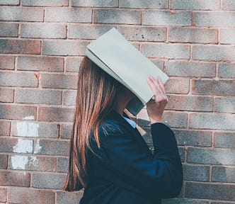 A girl with a book over her head. She’s standing next to a brick walla nd has long dirty blonde hair.