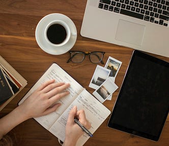 Overhead view of a wooden table used for composing an essay, on which lie an open laptop, a large electronic tablet, a small stack of books, some Polaroid photos, a pair of glasses (folded), black coffee in a cup and saucer, and an open business/student diary. A student’s hands, one of them holding a pen, partly obscure the handwriting on the diary’s pages.