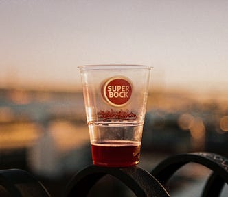 A half empty plastic cup of Portuguese beer sitting on a fence in evening light