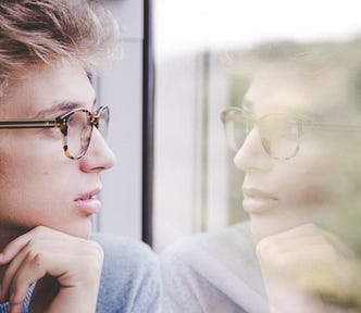 A young person with glasses looking from the window, putting their hand under their chin, and thinking.