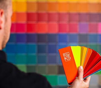 Someone facing away from the camera holding a fan of colour swatches. In front of them, away from the viewer, the wall is covered with out-of-focus squares of many colours.