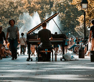 Person playing a baby grand piano in the park with people enjoying the sunshine as if there is nothing strange about a piano being there