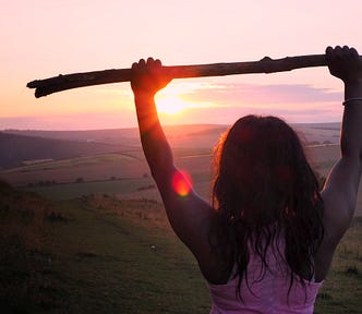 Woman looking over valley at sunrise holding stick in the air displaying an emotion of strength and conviction.