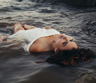 Glamorous woman in a white off the shoulder dress lying in beach water face up.