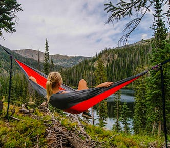 a photo that shows a young woman with a blond pony tail reclining in a hammock. We see her from the back as she looks out at the landscape of a distant lake, pine trees, and small mountains.