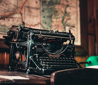 A gorgeous, antique black typewriter with a blank sheet of paper lying next to it, on a wooden desk.