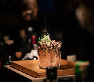 A mixed drink bubbling with a light green garnish in golden liquid and light — almost making it glow — sitting on a golden wooden board, elevated on a bartender’s counter but all else is dark around it except the silhouettes and shadows of liquor bottles.
