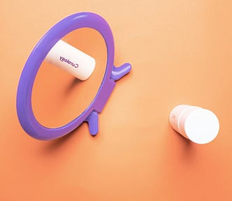 A vibrant purple mirror and skincare products are arranged against a warm orange background.