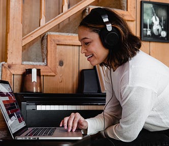 A woman with her headphones on, smiling and looking at the screen of the laptop.