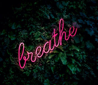 A neon light spelling out “Breathe” against a background of green foliage