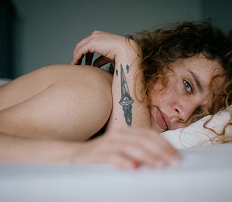 woman lies in bed, one arm up showing dagger tattoo