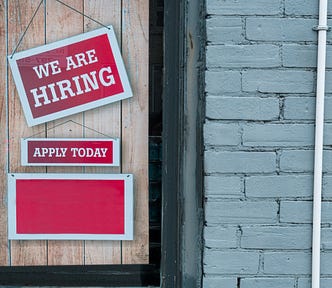 Brick wall with a window boarded up. On the window are the signs, “We are Hiring,” and “Apply Today.”
