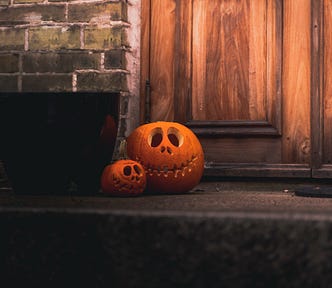 Two jack-o-lanterns sit on a porch in front of a wooden door.