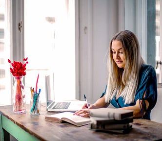 A female sits at their desk, writing in a journal with their laptop and pens nearby. #writing #journaling #writer