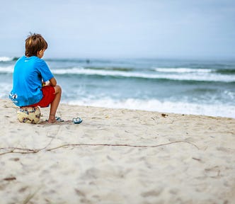 A little boy sits along staring at the waves along the shore.