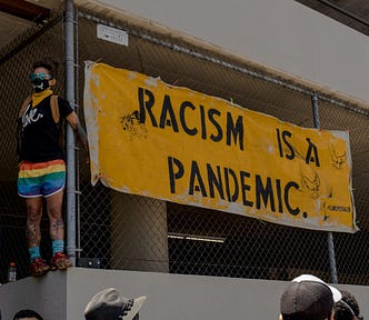 a person with rainbow shorts standing near a fence with the sign, “racism is a pandemic”