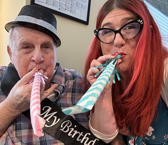 An elderly man in a hat and a middle-aged women with red hair and glasses, blowing party favors