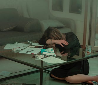 A woman sitting on the floor, burying her face into her arms and resting on a table.