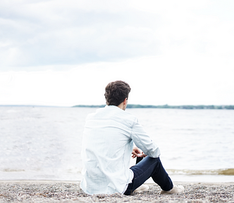 Man sitting on the beach looking at the water, casually dressed in white shirt, jeans, and tennis shoes.