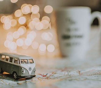 a toy VW campervan on a map on a table, in the background, out of focus, are fairy lights and a mug.