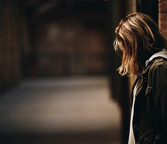 Unsplash image of a faceless woman with her head down, leaning against a brick wall. The background is a blurred alley.