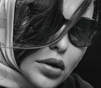A close-up in black and white of a beautiful woman whose face is mostly covered up in a head scarf, sunglasses, and long bangs as if to cover up her face.