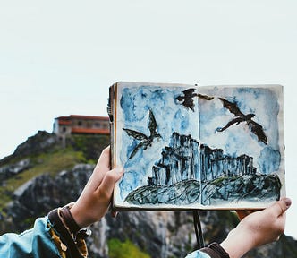 Image of a person’s arms and hands holding open a sketch book drawing of dragons soaring above a craggy cliff. In the background is an actual cliff with a low building at the top.