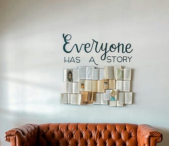 A brown leather couch sits below a wall with the phrase “Everyone has a story” and open books arranged in a grid.